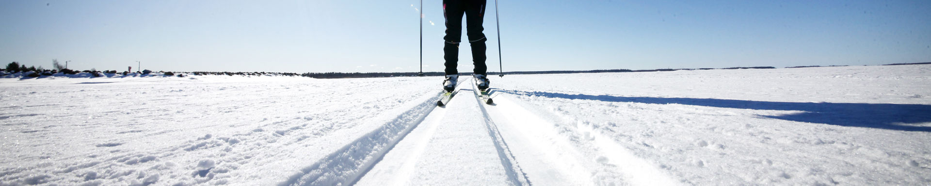 Hiihtolatu ja hiihtäjä. Aurinko paistaa sinisellä taivaalla.