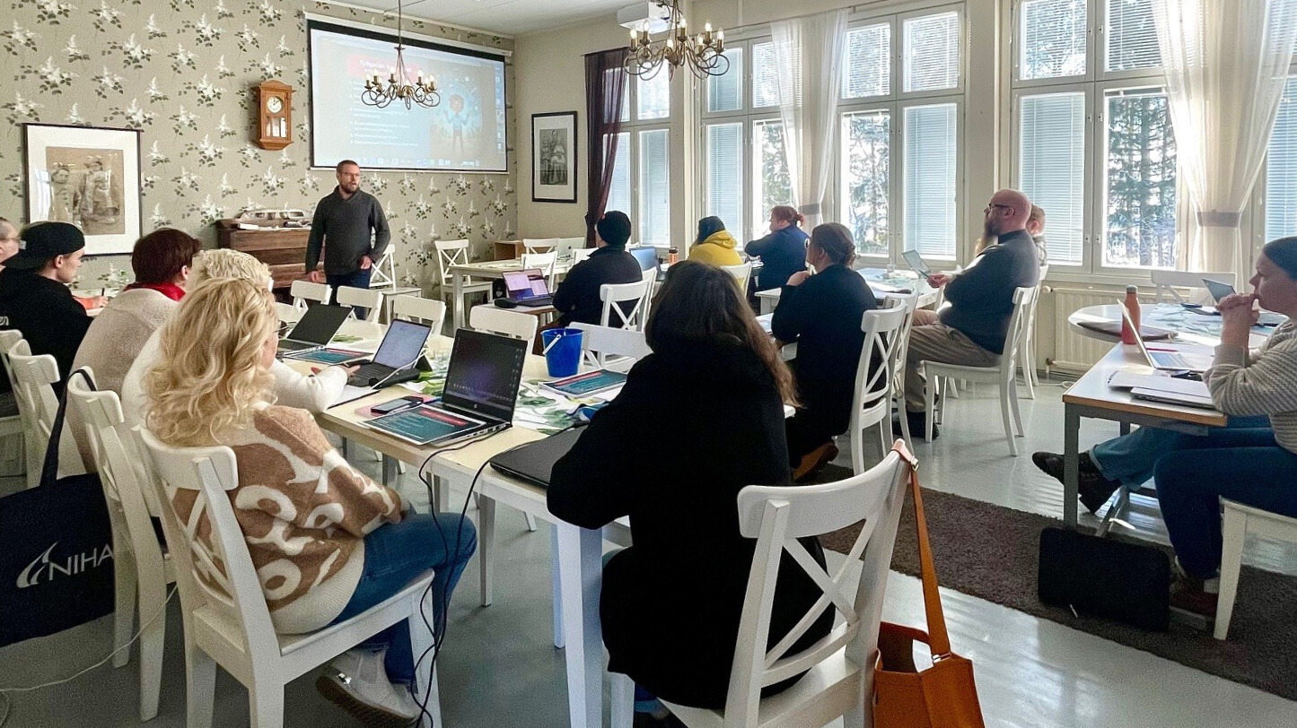 Kuvassa ihmisiä istumassa pöytien ääressä kuuntelemassa luennoitsijaa. 