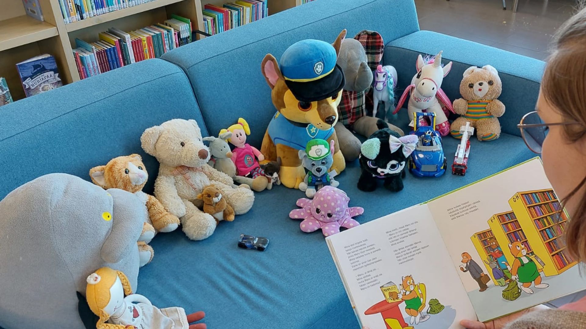 Pehmoleluja sohvalla. Etualalla satukirjaa pehmoleluille lukeva nainen.